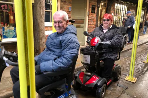 BBC / Naj Modak Man and women, both on mobility scooters on Shambles with scaffolding