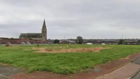 A large field with a brown patch of grass in the middle. There are houses and a church in the background. 