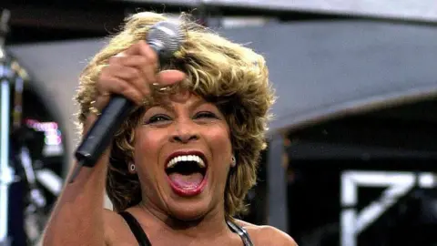 PA Media Tina Turner performing on stage. She is smiling with her mouth open as she smiles. She hold a microphone in one hand which is raised.
