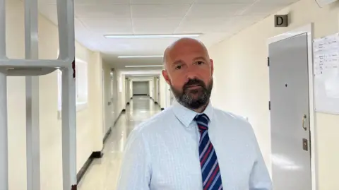 John De Carteret - A man with a bald head, and a black and grey beard looking at the camera. He's wearing a white and llight blue checked shirt, and a blue, pink, red and purple striped tie. He is standing next to some prison bars in a lonmg corridor of Guernsey's Les Nicolles prison. 