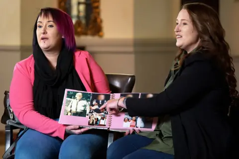 Two women are holding a photo album. The woman on the left is wearing blue jeans and a bright pink jacket. The one on the right is wearing a green blouse and a black jacket. 