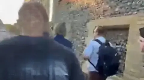 Facebook A man and a woman walk next to a brick building while being followed by a man wearing a shirt that says "Fork the System".