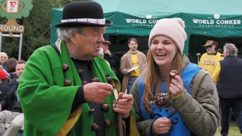 The World Conker Championships David Jakins and Kelci Banschbach are both holding their conkers. David has a face of disbelief and Kelci is laughing.