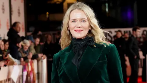Edith Bowman is standing on a red carpet and there are crowds standing behind a barrier in the background. She has blonde wavy hair and is wearing a dark green velvet blazer and red lipstick.