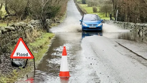 BBC A blue car driving through a small flood in a country road. There is a hazard sign warning of the flood and a cone in some standing water. 