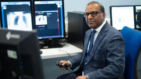 Leeds Teaching Hospitals NHS Trust Dr Fahmid Chowdhury is sitting on the right. He is sitting on a blue seat and is holding a remote. In front of him is a screen with two chest X-rays on it