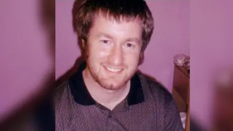 Family Photo Thomas Reeve smiling at the camera has ginger hair and beard with blue eyes and is wearing a purple shirt.
