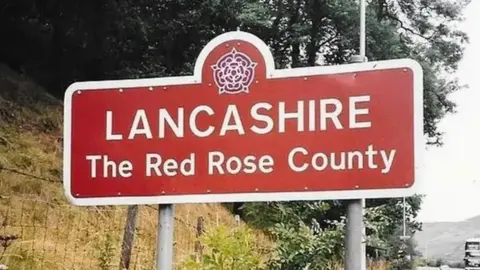 A red road sign with the words Lancashire, The Red Rose County on it in white letters.