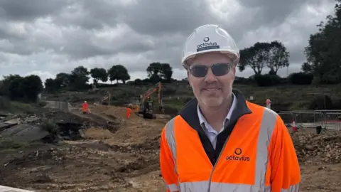 Chris Hudson in high-vis clothing, hard hat and sunglasses looking into camera with a building site behind