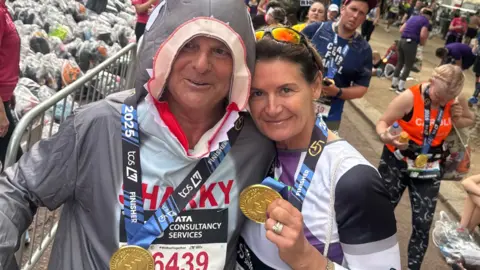 Handout Shorting and her husband posing for a picture at the finish line of the London Marathon 2025, with gold medals around their neck. They are both in fancy dress - he is wearing a shark onesie and she is dressed as Buzz Lightyear. They are surrounded by other runners and a metal barrier.