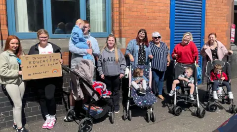 A group of protesters stand a line outside the Education Authority's red brick HQ in Belfast. Many of them are mothers wheeling children in buggies. One young girl is holiding a cardboard sign which reads: "I deserve an education like my friends."