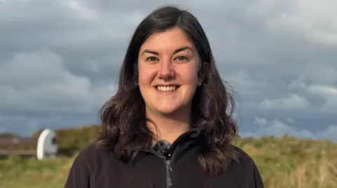 Niamh McDevitt pictured with shoulder length dark brown hair smiling at the camera with teeth. She has a septum piercing. She is wearing a black zip up fleece with the Alderney Wildlife Trust logo on.