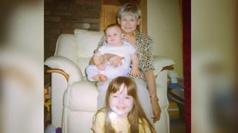 Family photo An older woman with short blonde hair, sat on a cream armchair, wearing a leopard print blouse. On her lap is a blonde baby dressed all in white, and in front of them, on the floor, a young girl with long hair, wearing a yellow t-shirt, is sat. They are all smiling and looking at the camera.