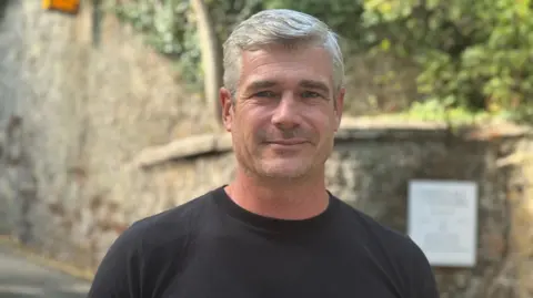 Harry Trower has silver hair is pictured in a black T-shirt. The walls of Chateau la Chaire's gardens are visible behind him with some foliage.
