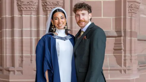 Fotografía de la Universidad John Moores de Liverpool: La novia y graduada Charnele Washbourne, con vestido de novia y velo, y su toga de graduación, junto a su prometido Bradley Natt, de cabello castaño y barba, que viste un traje de boda gris oscuro. Ambos se encuentran dentro de la Catedral Anglicana de Liverpool y sonríen.