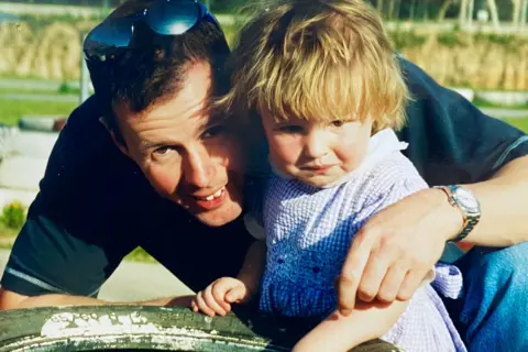 Hollie McRae Colin McRae and Hollie as a toddler. They are both positioned behind a large tyre that has been painted white. Colin is wearing a dark shirt and jeans, and has sunglasses on his head. Hollie is wearing a blue and white gingham dress with a white collar. The background is slightly blurred, but appears to be an outdoor setting with trees and greenery.