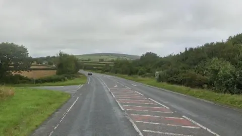 Google Street View image of the A388 near St Dominick in Cornwall. A car is driving along the road in the distance. A t-junction on the left-hand side. Trees, bushes and fields are along both sides of the road. It is a cloudy day.