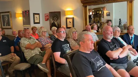 A group of people are sitting indoors in a pub, looking at a screen which is out of shot. The walls of the room have various paintings and mirrors on them.