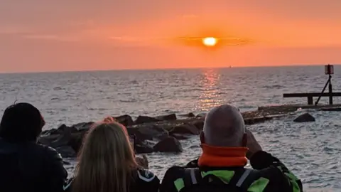 A group of three people viewed from behind look out at a vibrant orange and pink sunset over a calm sea, with rocks and a small wooden pier structure visible on the right.