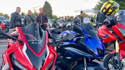 The motorbikes, two red and one blue. The red motorbike on the left has a sticker of the windshield reading ‘In memory of Josh. Forever 21.’ Three people in bike leathers stand behind the bike.