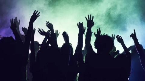 Dancers in silhouette amid lights and smoke raise their hands in the air 