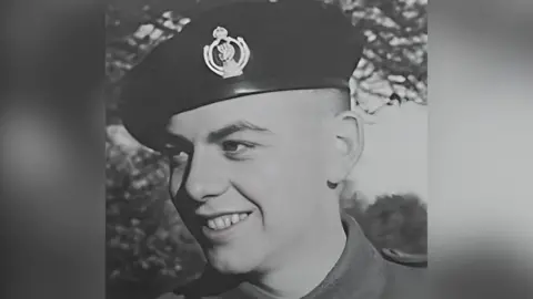 A black and white photo of Trp Ashworth in his military uniform, he is smiling. There are trees in the background. 