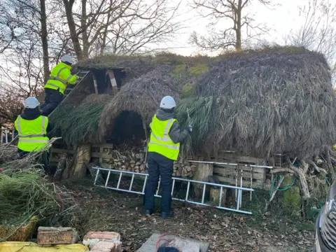 A small basic stone building with a messy, work-in-progress thatched roof. Straw is sticking out in all directions while three thatchers in hi-vis and white hardhats work on the process. A ladder sits at the bottom of the stone wall and one is leant on the left side with a worker at the top.