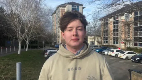 Sion looking at the camera. A car park and the university building are behind him. He is wearing a beige hoodie. 