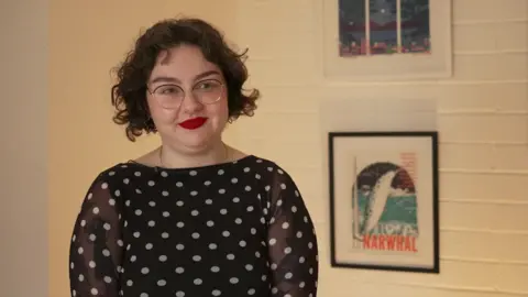 There is a picture of a lady with a black polka-dot top in an art gallery with pictures on the wall behind her.