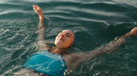 A woman wearing a blue tankini and a blue swimming cap floats in the sea with her arms outstretched behind her.