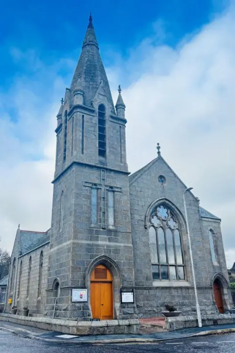 Strichen Church, with a tall spire, and a blue cloudy sky.
