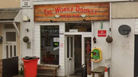 Google A micropub with a brown sign above the door which reads Wonky Donkey, micro pub. The pub has a white front door and a large window to the left hand side.