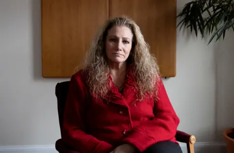 Marlean Ames ints in a chair weraring a red blazer  and black pants with her hair falling over her shoulders