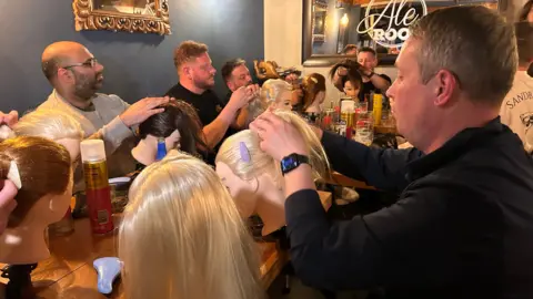 A group of men sitting around a long table. In front of them are dolls' heads attached to the tables, each with different colour hair. There is hairspray, brushes and clips on the table and the dads are playing with the hair.