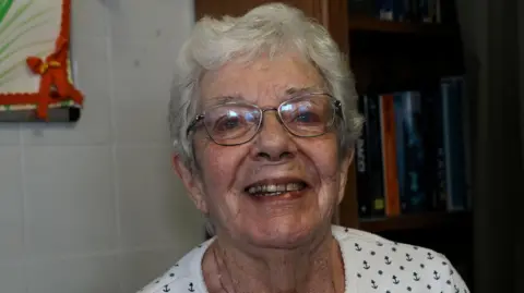 A head and shoulders image of a woman wearing glasses and a white top. She is looking into the camera while smiling. 