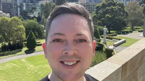 Jamie Welch A white man with brown hair is smiling in a selfie while standing in front of a large garden 