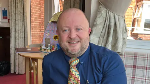 Sid Martin, Business Manager at Milward House, sits on sofa in the lounge with a dolls house behind him