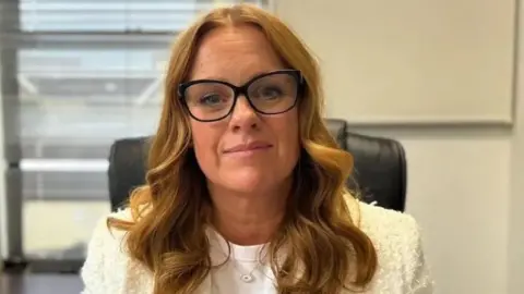 BBC Claire Christian, a woman with red/brown hair, she wears large glasses and a white blazer and looks at the camera.