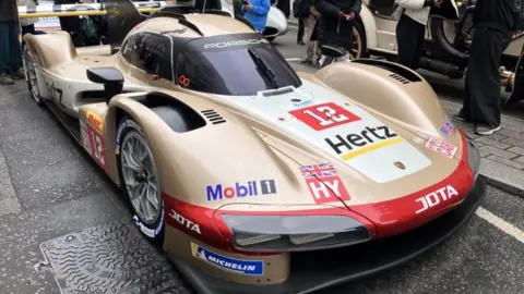 Asad Ahmad/BBC A gold Porsche 963 hypercar, covered in advertising stickers for the likes of Michelin and Hertz, parked on Pall Mall