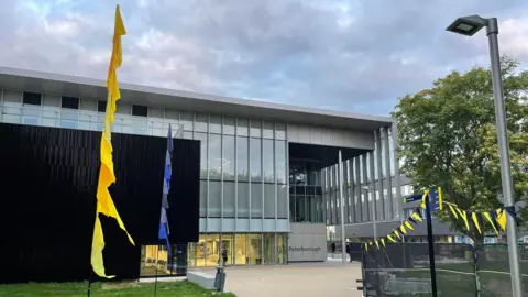 Ben Schofield/BBC Front of ARU Peterborough, a glass-fronted building where there is a path leading to it flanked by yellow and blue bunting.