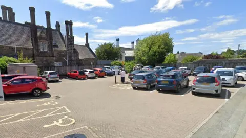 A car park is seen with a number of vehicles and buildings in the background on a sunny day