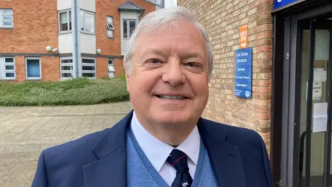 A man is standing outside a building, wearing a dark blue suit, blue jumper, white shirt and navy tie.