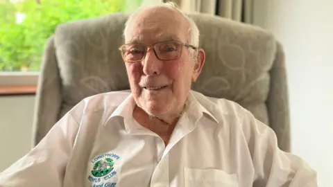 Richard is sitting in a chair and wearing a white shirt with the logo of Longthorpe Bowls Club on it. He is  smiling for the camera.