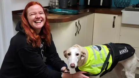 Emily Devine who has red hair, is laughing as she sits with her dog - a white lurcher, who is wearing a hi-vis jacket