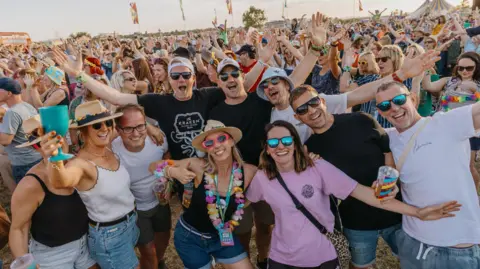 Andrew Whitton A group of ten friends smile and have their arms linked as they pose for a group shot at the Wychwood Festival. In the background a large crowd is visible. It is a sunny day and many of the people are wearing sunglasses