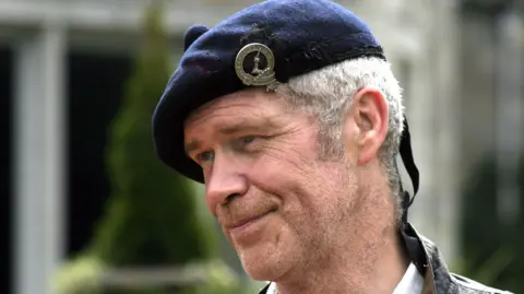 Sandy Morton, smiling in close-up in a tartan hat. He has grey hair.