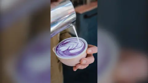 A hand holds coffee cup. Above it, milk is pouring from a jug to create foam art in a purple drink.