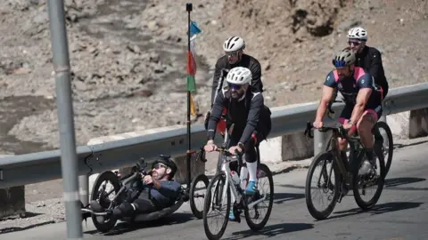 Ben Large A handcyclist lies low to the ground on a mountain road, travelling alongside a group of upright cyclists during the expedition.