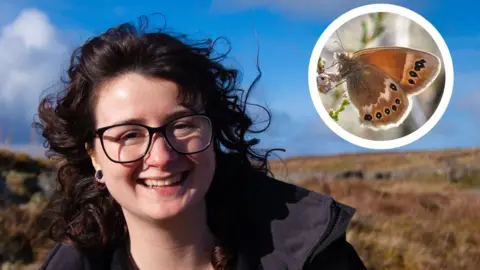 Georgina Paul smiles at the camera. She has curly dark hair and black framed glasses. She stands outdoors in a grassy, heath on a sunny day. In the top corner, a circular inset shows a close-up of a Large Heath butterfly - it is brown and orange with small black eye spots on its wings.