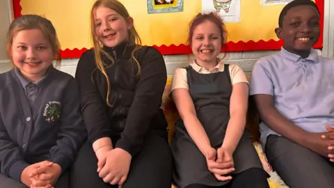 Simon Thake/BBC Three young girls and a young boy sit together with their arms folded on their laps. They are all smiling and wearing a variety of different school uniforms in blue, grey and black.